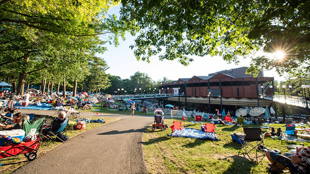 A look down the walkway of the Saratoga Performing Arts Center lawn with people sitting in lawn chairs and the sun peeking through a green chair to the left