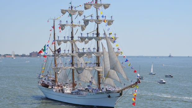 A Tall Ship fro Sail 4th celebration floats on the water. The mast are decorated with the flags of various nations and people stand from the yards.