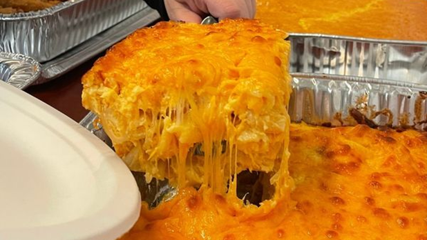 A person scoops a heaping helping of creamy mac and cheese from a tray at Allie B's Cozy Kitchen