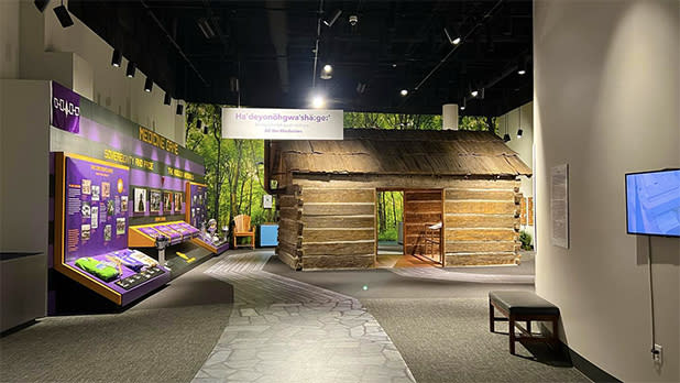 All The Medicines exhibit at the Seneca-Iroquois National Museum. Purple signage with information on the left and a log cabin on the right.