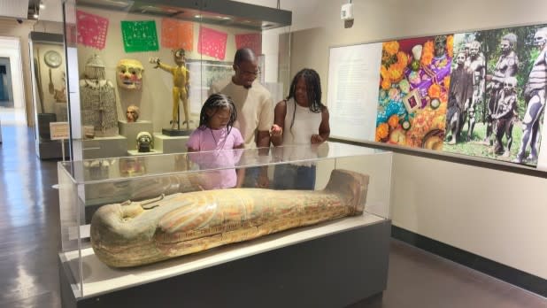 A family of three observes an ancient Egyptian sarcophagus at the Buffalo Museum of Science. Colorful artifacts and a vibrant cultural exhibit are displayed in the background.