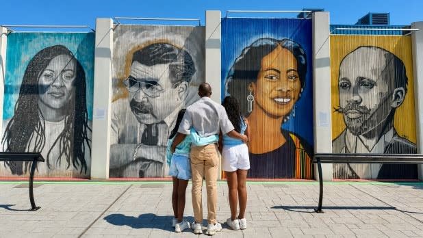 A family of three embraces while admiring the Freedom Wall in Buffalo, a collection of large, colorful murals featuring historical figures in a public outdoor space, evoking a sense of reflection.