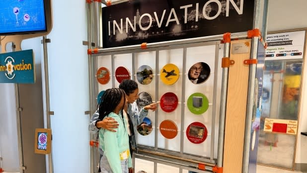 A woman and a young girl stand in front of an "Innovation" exhibit at Explore & More - Ralph C. Wilson, Jr Children’s Museum. They engage with colorful circular panels featuring images and text, conveying curiosity and learning.