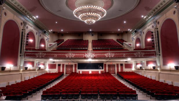Interior of an opulent theater, red chairs, lit chandelier, and balcony