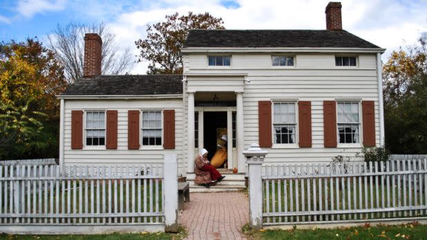 A white colonial-style house with brown shutters, a brick path, and a white picket fence. A person in historical dress sits on the steps, surrounded by autumn trees.