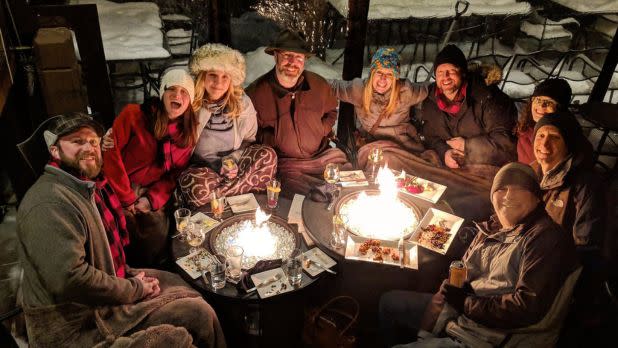 Group of people in winter clothing sitting around two fire pits with food