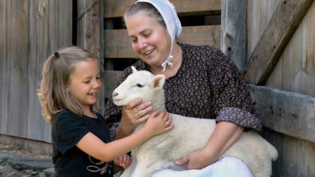A woman and a young girl joyfully pet a lamb, sitting by a wooden barn. The woman wears a bonnet, and the scene conveys warmth and rural charm.