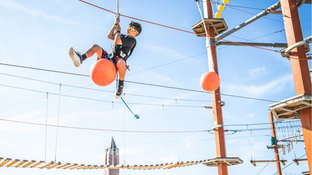 A child wearing sunglasses joyfully navigates a high ropes course, clinging to a rope beside an orange ball under a bright blue sky.