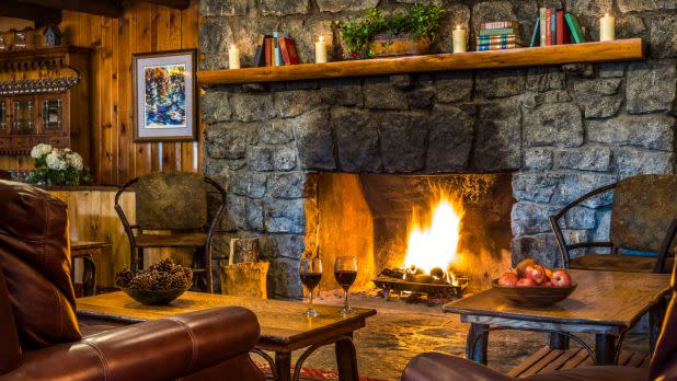Cozy living room with a stone fireplace emitting a warm glow. Shelves hold books and candles. Two wine glasses and apples on a wooden table suggest relaxation.