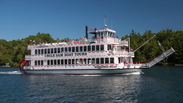 Tour boat with red trim on a lake under a clear blue sky, surrounded by lush green trees. The boat's upper deck is filled with people enjoying the sunny day.