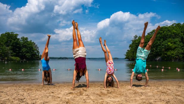 Four people perform handstands on a sandy beach, with a serene lake and lush trees in the background. The sky is bright blue with fluffy clouds.