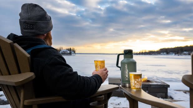 A man wearing a grey beanie and jacket sits in a plastic Adirondack chair, holding a glass of beer and looking out over a frozen, snow-covered lake at sunset. A large green thermos and another glass of beer rest on the chair's armrest beside a small, lit fire pit.