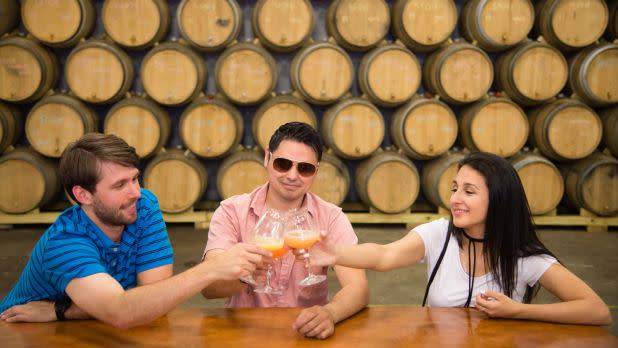Three people—two men and one woman—sit at a wooden table in a brewery, toasting each other with wine glasses containing an orange-colored beverage, set against a backdrop of stacked wooden fermentation barrels.