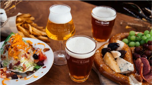 A wooden table with three glasses of beer, a leafy salad topped with shredded carrots and olives, a large pretzel with cheese and charcuterie, and a bunch of grapes. The setting conveys a cozy, inviting atmosphere.