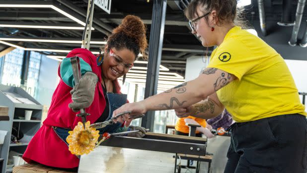 Two women collaborate on a glassblowing project. One, wearing a red jacket, shapes molten glass, while the other, in a yellow shirt, assists, both smiling.