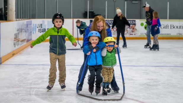 An adult woman helps two little kids ice skate on an indoor ice rink, an older child ice skates unassisted next to them