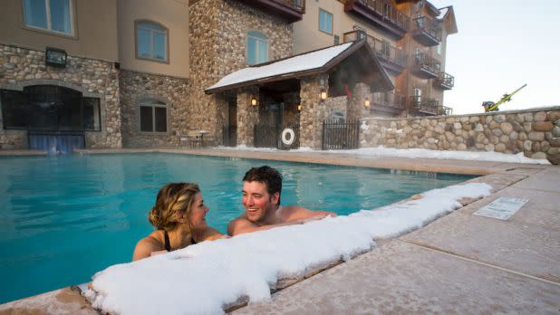 A couple smiles and relaxes in a heated outdoor pool surrounded by snow, next to a rustic stone building. The atmosphere is warm and cheerful.