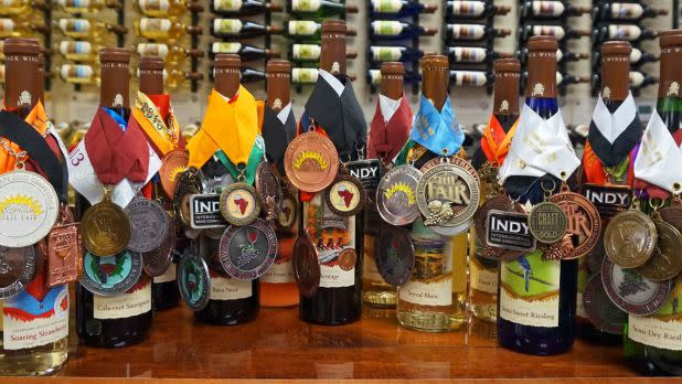 A row of wine bottles are adorned with numerous competition medals, showcasing their award-winning status. The background displays more wine bottles on shelves.