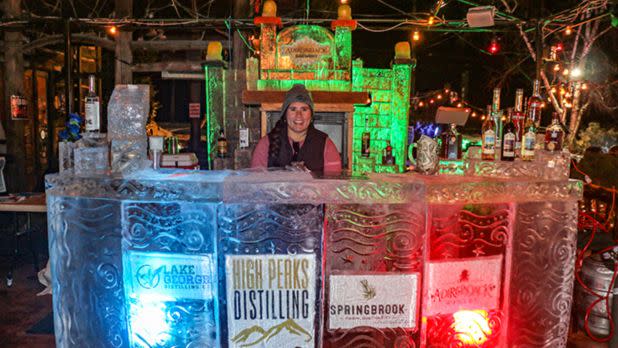 A person in winter attire stands behind an ice bar, featuring glowing bottles and colorful, illuminated logos. The scene is festive and lively.