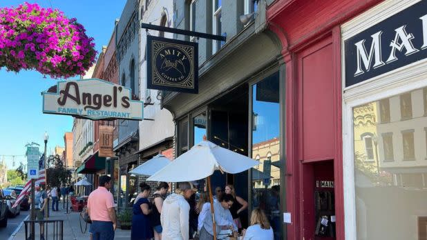 People gather outside bustling street cafes with umbrellas, signs visible for Angel's Family Restaurant, in a vibrant downtown scene. Bright flowers hang above.