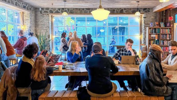 People sit at a cozy cafe with books and laptops on a large wooden table. A large window reveals an outside view, creating a warm, inviting atmosphere.