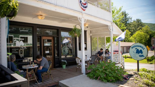 Outdoor cafe scene with people seated at small tables on a porch. The sign reads "Mornings." A sunny day highlights a cozy, relaxed atmosphere.