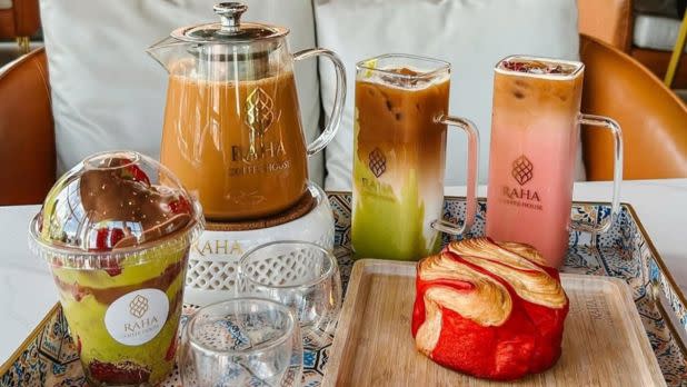 Assortment of drinks and pastries on a tray. Iced teas in tall glasses, one pink, one with milk and green tea. A vibrant red pastry is in front, creating a cozy, inviting feel.