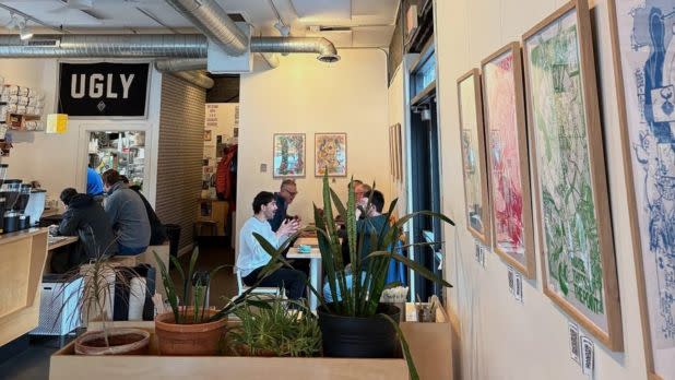 A cozy café with modern decor, featuring framed art on the walls, potted plants, and several people engaged in conversation at tables, with a casual, friendly atmosphere.