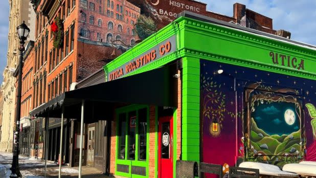 A vibrant street scene shows a bright green building with "Utica Roasting Co" signage. Artistic murals adorn the side, depicting a nighttime landscape. Nearby, a historic brick building features a vintage-style mural. The scene conveys a lively and artistic urban atmosphere under clear, sunny skies.