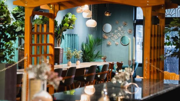 Cozy restaurant interior with warm lighting. A long table with chairs, green plants, and wall decor saying "Good Vibes Only" create a welcoming atmosphere.