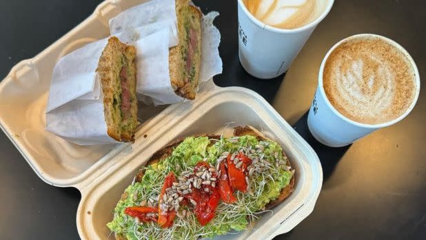Open takeout box with a sandwich and avocado toast topped with sprouts and red peppers. Two cups of frothy coffee sit beside on a dark surface. Cozy and appetizing.