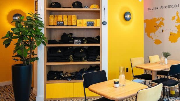 Bright cafe interior with vibrant yellow walls, cozy wooden tables, and chairs. Shelves display black merchandise and coffee bags. A potted plant adds greenery.