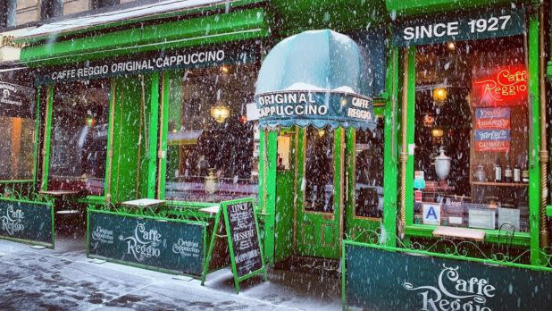Snow falls gently outside a vibrant green café with a retro awning. Soft lights inside create a warm, inviting atmosphere. Sign reads: "Since 1927."