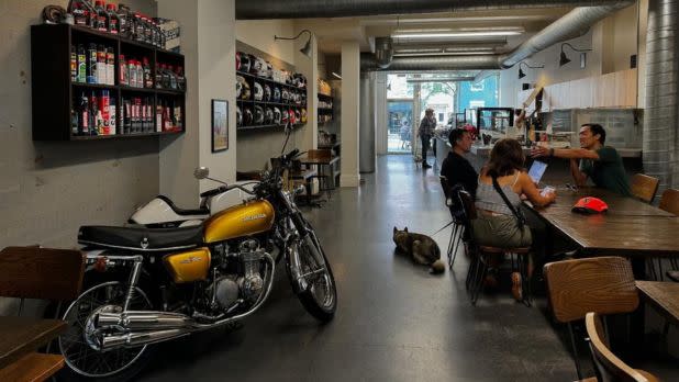 Interior of a stylish café with a vintage motorcycle on display. People sit at wooden tables, conversing, while a dog rests nearby. Warm, inviting atmosphere.