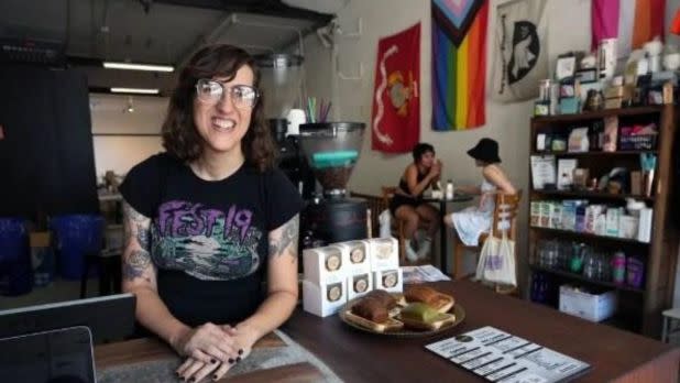 A smiling barista stands at a cafe counter with pastries. Pride and other flags hang on the wall. Behind, two women sit chatting. Cozy and welcoming vibe.
