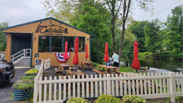 A cozy coffee lodge with Catskills signage, outdoor seating, red umbrellas, a white picket fence, lush greenery, and a serene riverside setting.