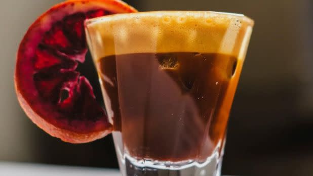 A glass of espresso topped with frothy crema, garnished with a deep red slice of blood orange. The image conveys a vibrant, refreshing mood.