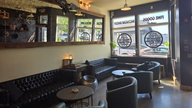 Cozy café with leather sofas, round tables, and large windows. Mirrors reflect the warm, inviting space. Ambient lighting and outdoor view.