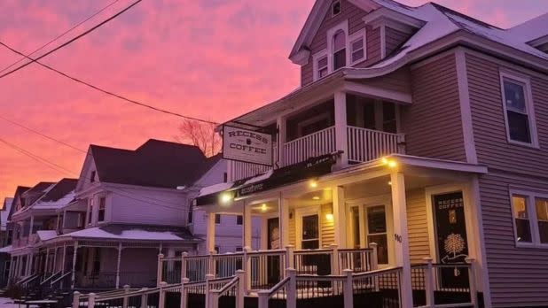 Historic café in a large, two-story house with a wraparound porch, warmly lit at dusk. The sky is a vibrant pink and orange, creating a cozy atmosphere.