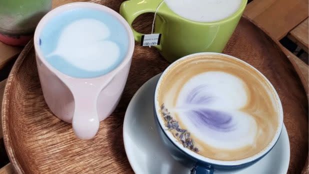 Three mugs of latte on a wooden tray: a pink mug with blue latte art, a green mug with tea, and a blue mug featuring lavender and heart art.