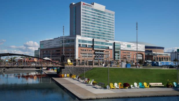 A tall modern hotel overlooks a waterfront with colorful chairs on the grass. A clear blue sky adds a serene, inviting atmosphere to the scene.