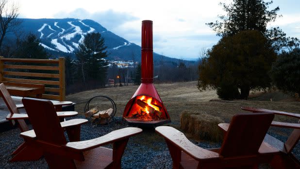 Cozy outdoor fire pit with red chimney, surrounded by wooden chairs. A snowy mountain looms in the background, creating a serene, wintery atmosphere.