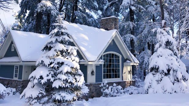 Cozy blue cottage surrounded by snow-laden pine trees, evoking a serene winter scene. The roof and ground are covered in thick, glistening snow.