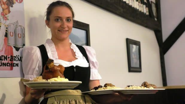 A smiling server in traditional attire holds plates of hearty food, including roast meat and mashed potatoes, in a cozy, warmly lit restaurant.