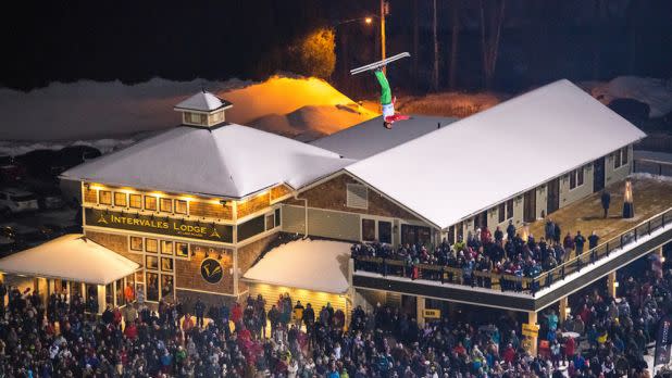 A large crowd gathers at night outside a snow-covered lodge, intently watching a skier mid-air, performing a flip against a dark winter backdrop.