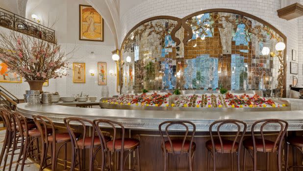 Elegant restaurant interior with an ornate bar adorned with colorful flowers. Curved wooden chairs, mosaic mirrors, and soft lighting create a cozy, welcoming ambiance.