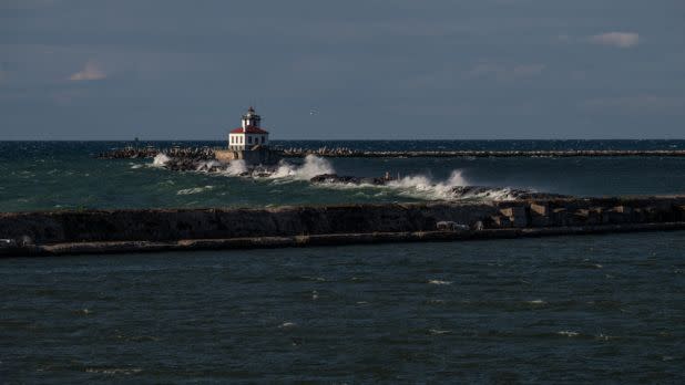 Large, white-capped waves crash violently against a long stone breakwater protecting a white and red-roofed lighthouse on a stormy, overcast day.