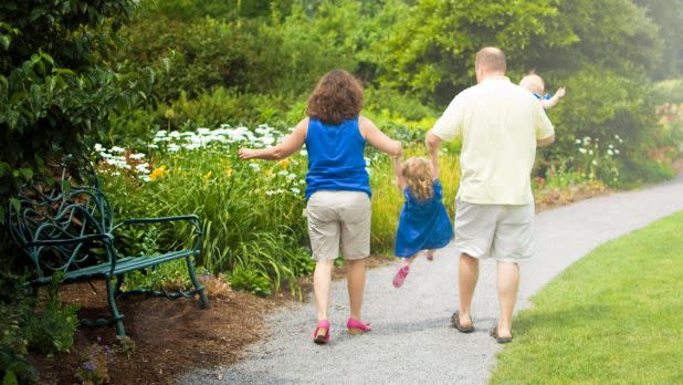 A family of four seen from behind as they walk down a gravel path winding through a lush botanical garden filled with green bushes and white flowers.