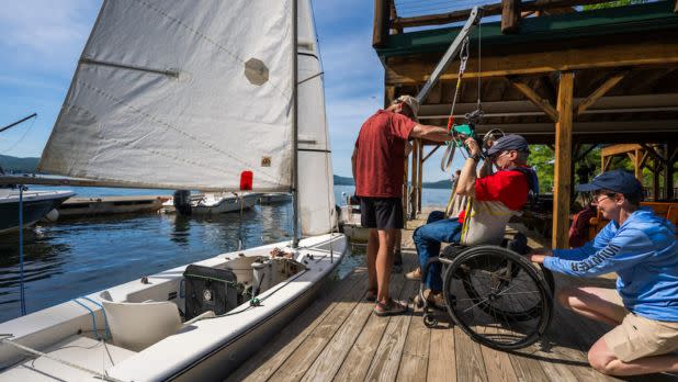 Volunteers on a wooden dock use a mechanical hoyer lift to assist a man in a wheelchair as he prepares to transfer into an adaptive sailboat on a sunny day.