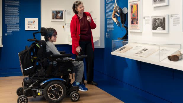 A young individual in a power wheelchair and a woman in a red cardigan smile while exploring a museum exhibit with framed photographs and glass display cases against vibrant blue walls.